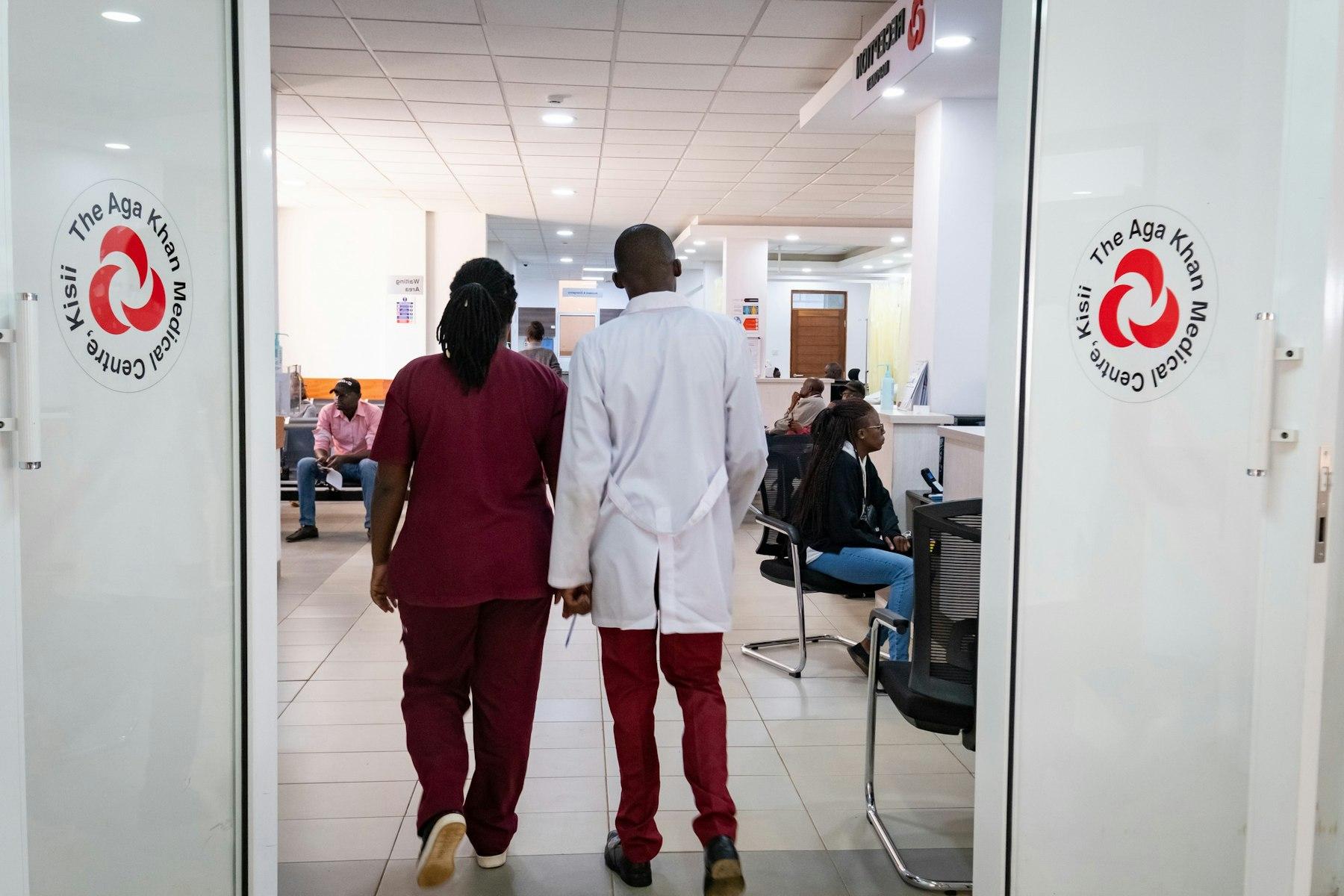 Medical staff walk through the waiting room past a door with a logo showing The Aga Khan Medical Centre, Kisii.