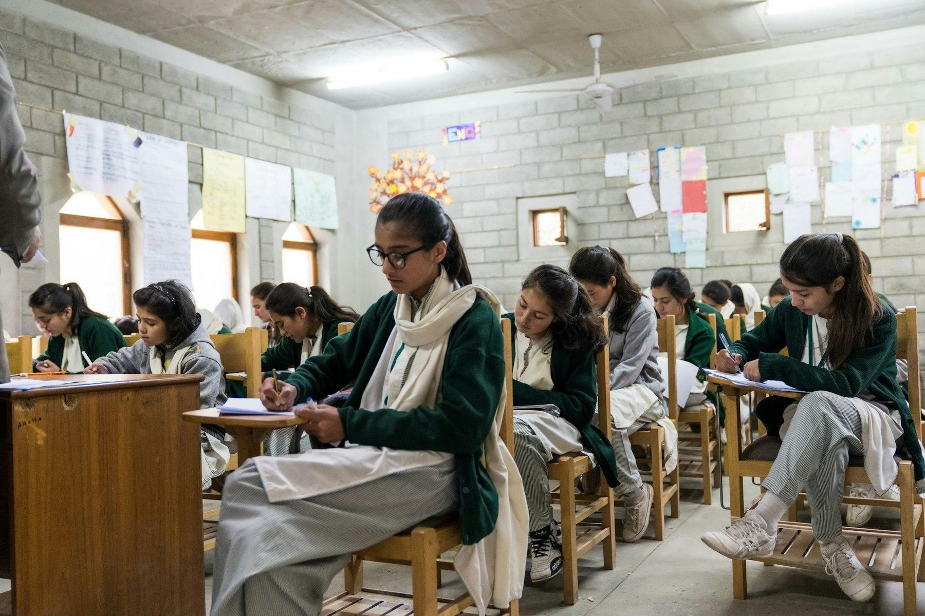 Girls in green and grey uniforms sit at desks concentrating on their writing.