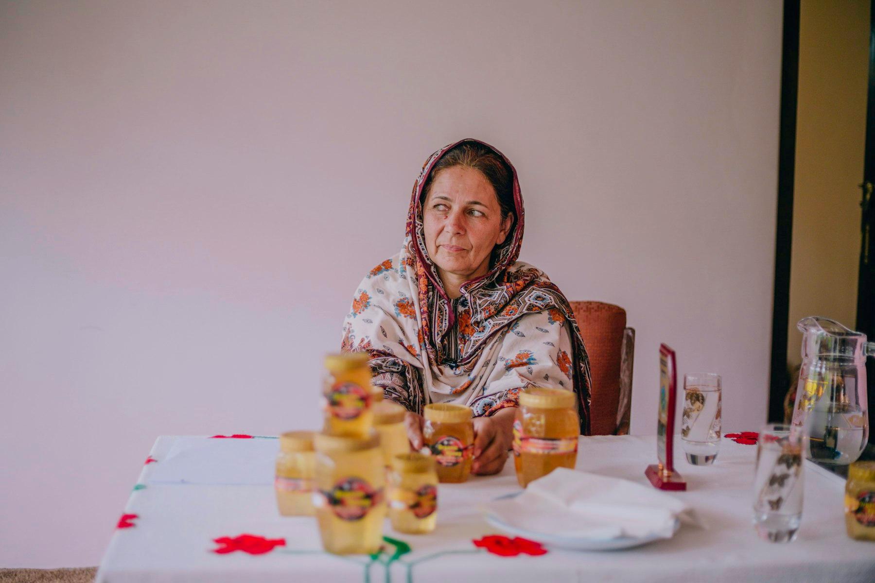 A woman sits at a kitchen table with several jars of honey.