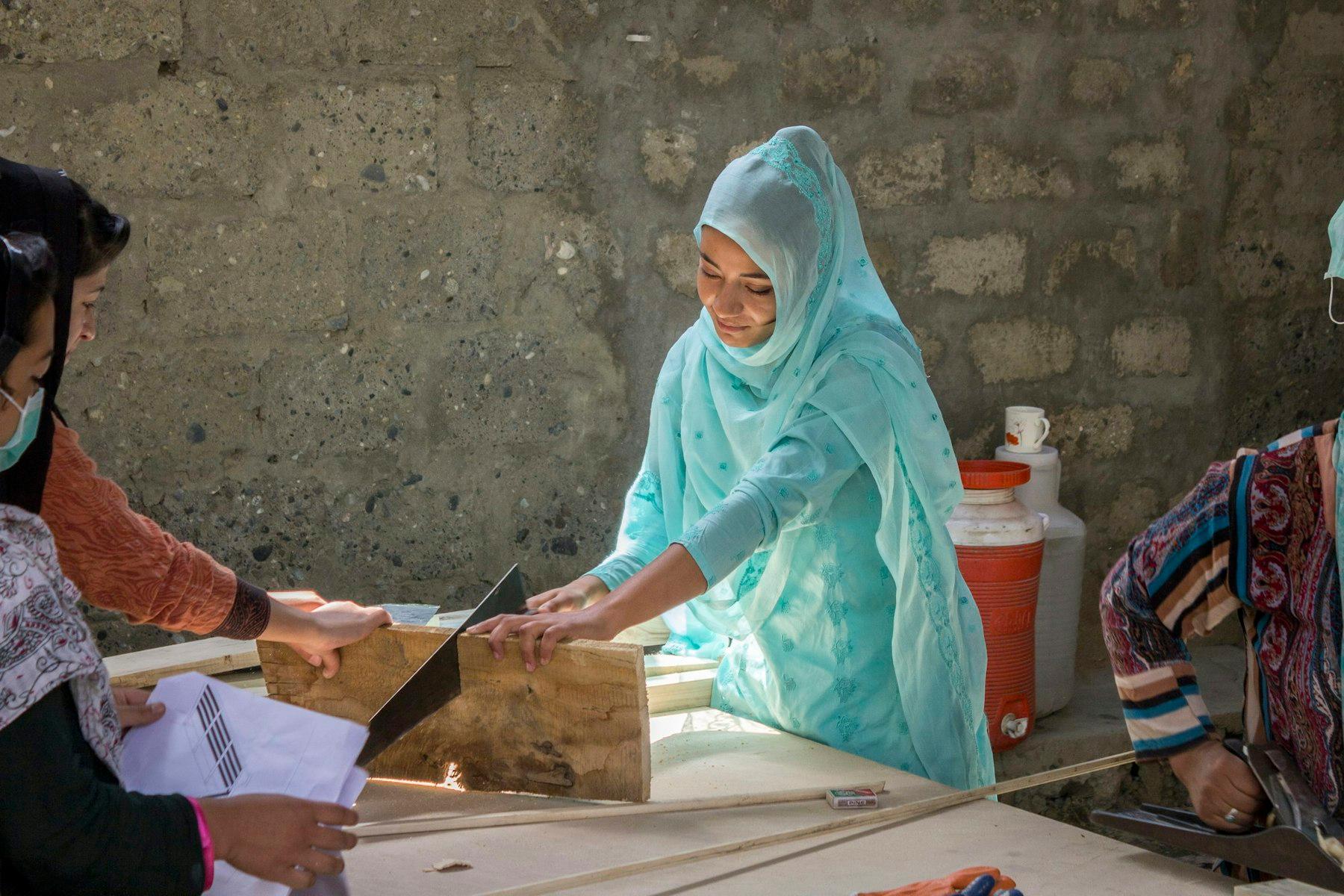 A woman saws through a plank that another woman holds, while a third holds a plan.