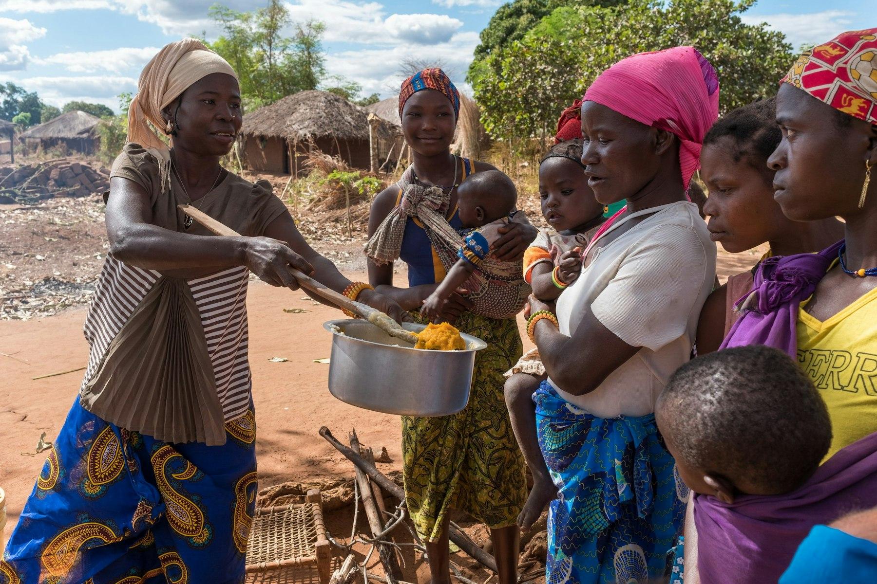 A woman holds out a saucepan of porridge to show a group of women with their babies.