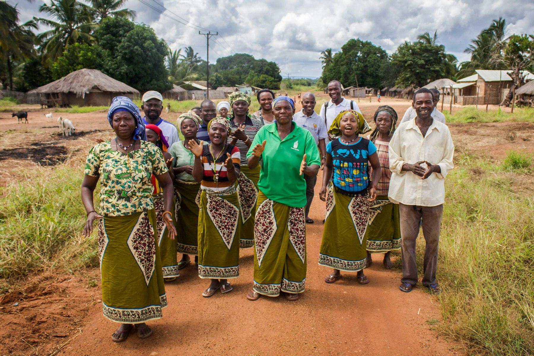 Homens e mulheres sorriem e aplaudem, num trilho com campos, cabras e casas à sua volta.