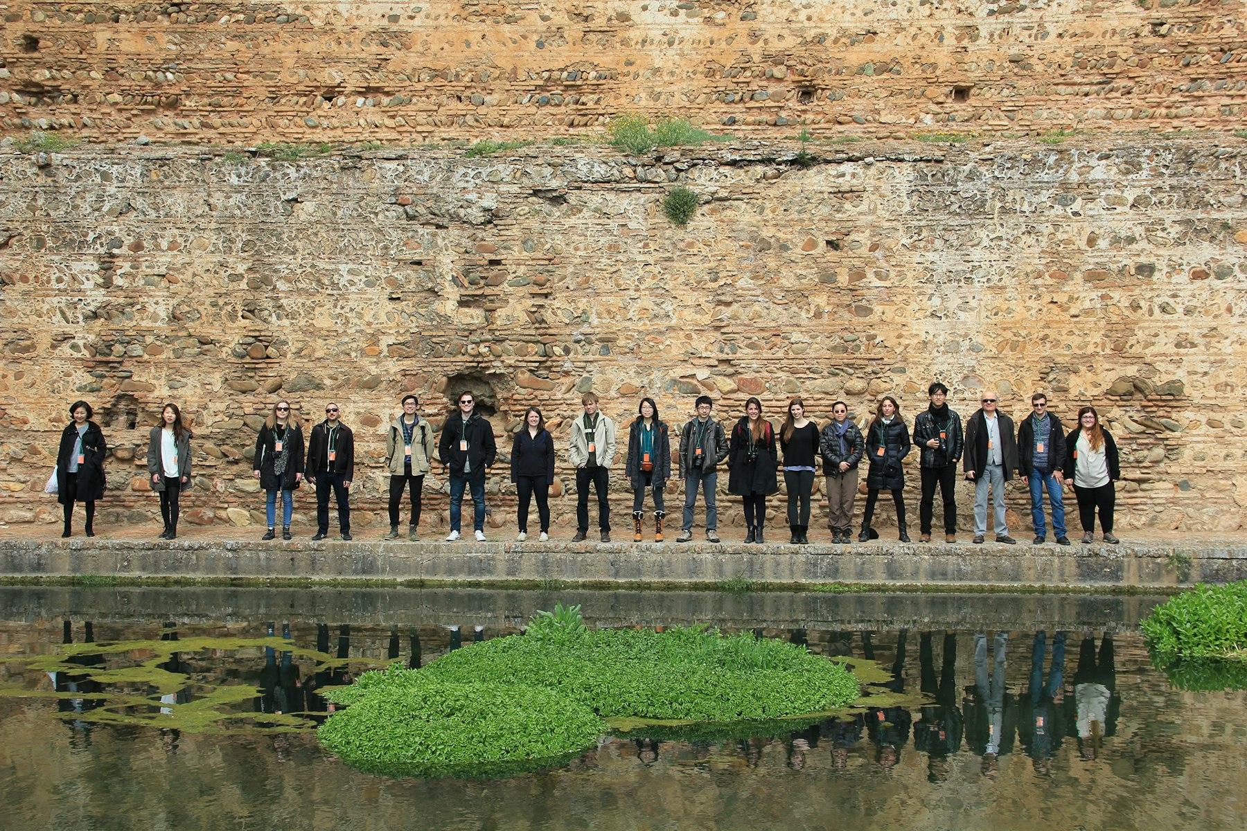 A group of visitors stand near a tall wall, reflected in the water by their feet.