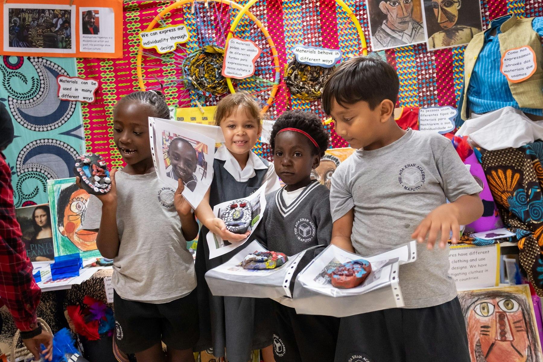 Four children, standing by a brightly decorated wall, show their craft projects.