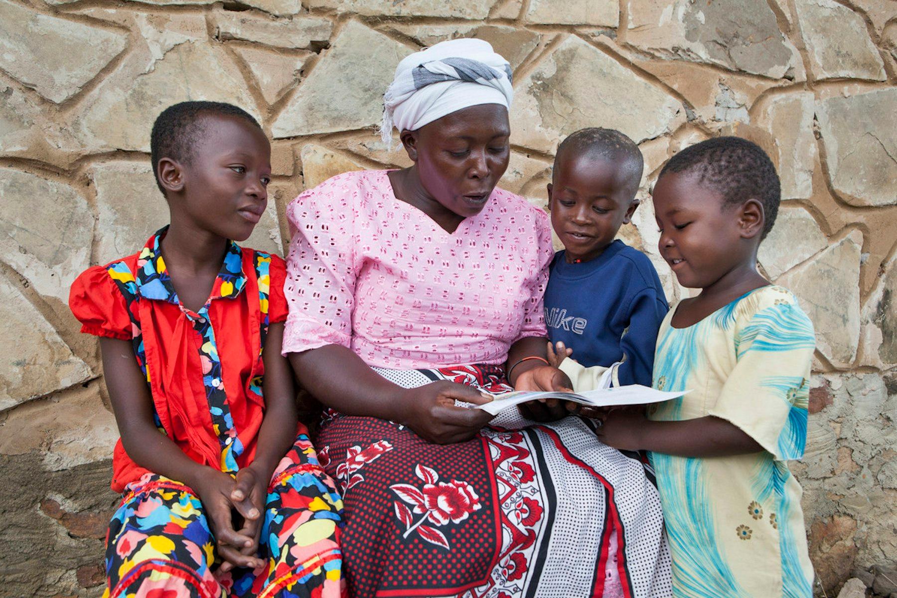 A woman sits outdoors with three children. They look at a book together.