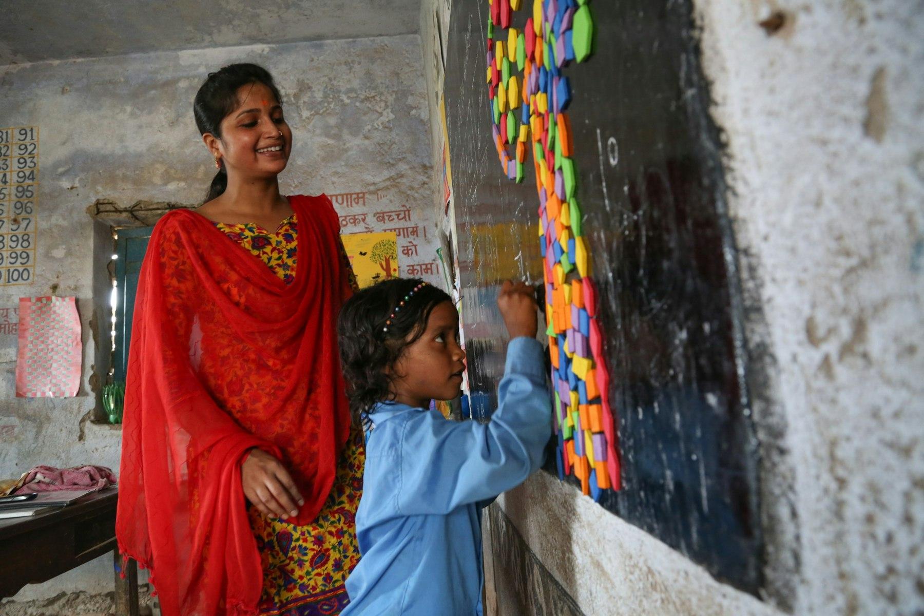 A child writes on a blackboard while an adult watches approvingly.