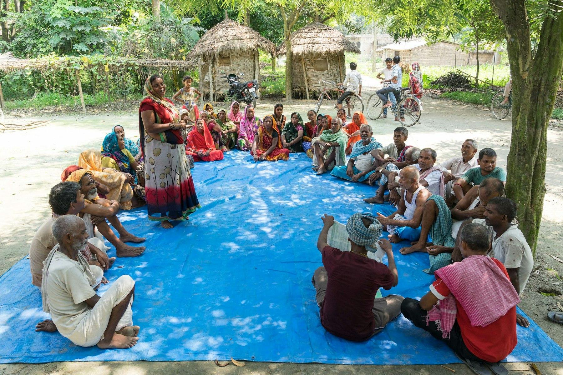 A group of villagers sit in a circle on a blue mat. A woman stands and talks while a man holds up a document.