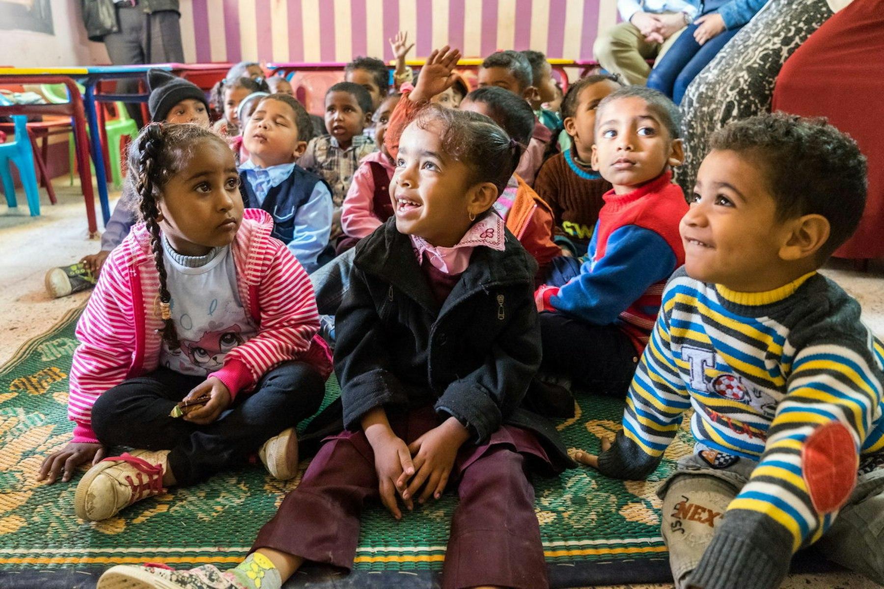 Des enfants sont assis sur un tapis, et certains lèvent la main pour répondre à une question.