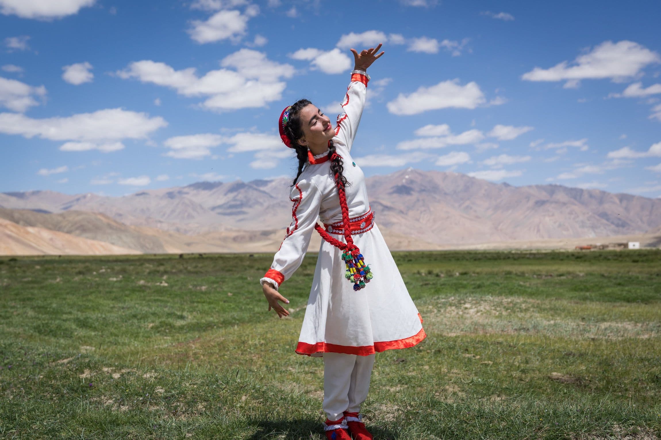 A woman in a white dress with red trim dances on grass with mountains in the background.